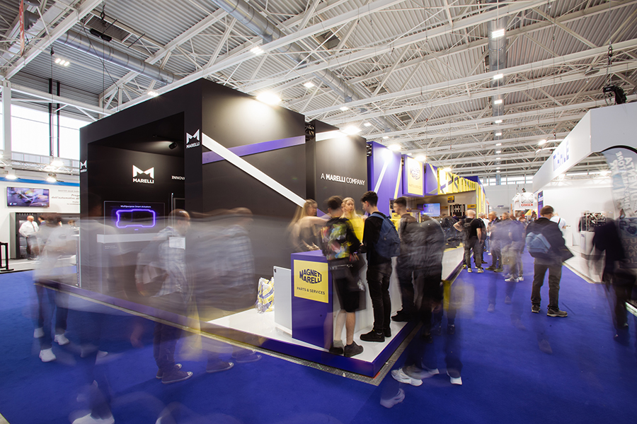 Side view of the Magneti Marelli Parts & Services stand at Autopromotec 2025: in the foreground the Marelli tunnel with frontier technologies, in the background the yellow and blue company logo and visitors in the adjacent aisle.